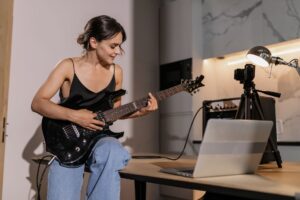 Musician girl playing electric guitar indoors with camera setup for recording, in a modern setting for online audience.