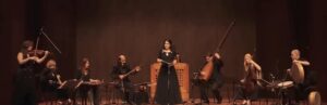 Chamber orchestra with instruments from baroque era and perisan instruments and singer on a stage performing Bach & Khayyam