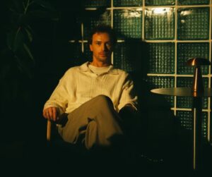 A male musician sitting ona chair looking at the camer in the dimly lighted room. Cascade by Alex Solely, Album review.