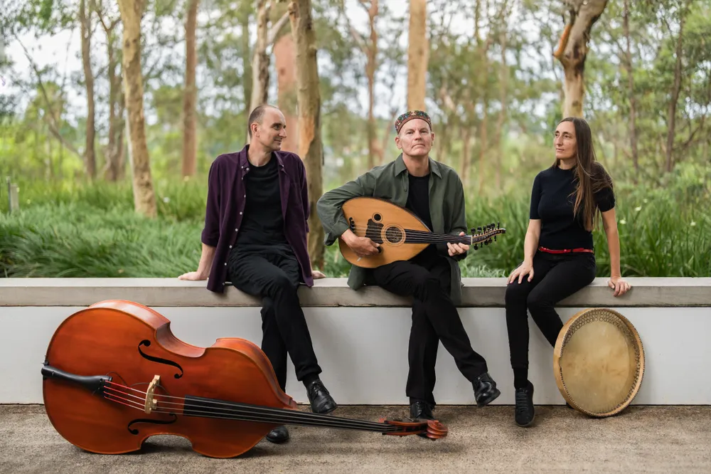 Music band East of West, Two male and female musician sitting ona curb. A double bass on the ground, a oud in the hand a musician and a percission instrument on the right side of the frame.