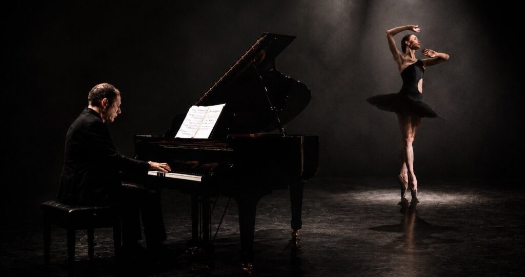 A pianist behind a black grand piano and a ballerina with dark clothes on a dimmly lighted stage.