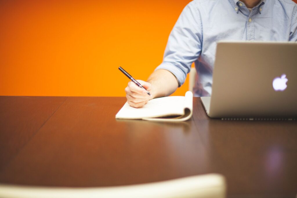 Adult writing notes in a notebook beside a laptop on a wooden desk with orange background