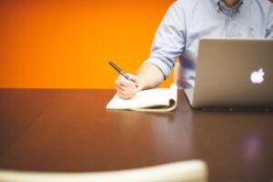 Adult writing notes in a notebook beside a laptop on a wooden desk with orange background