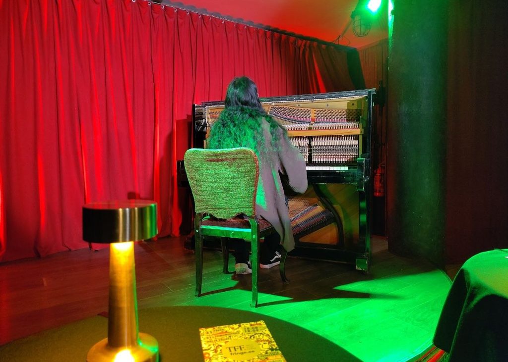 Pianist on the stage performing on an open upright piano with red curtains in the back and green light on the stage.