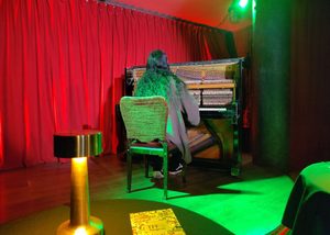 Pianist on the stage performing on an open upright piano with red curtains in the back and green light on the stage.