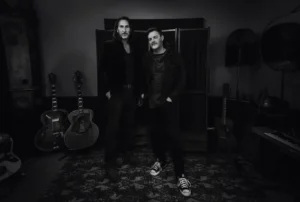 Black and white picture of two male musicians standing in a music studoi with guitars behind them.