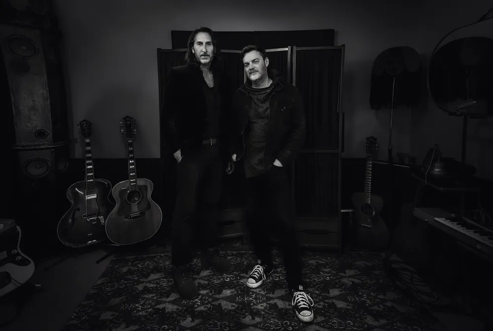 Black and white picture of two male musicians standing in a music studoi with guitars behind them.