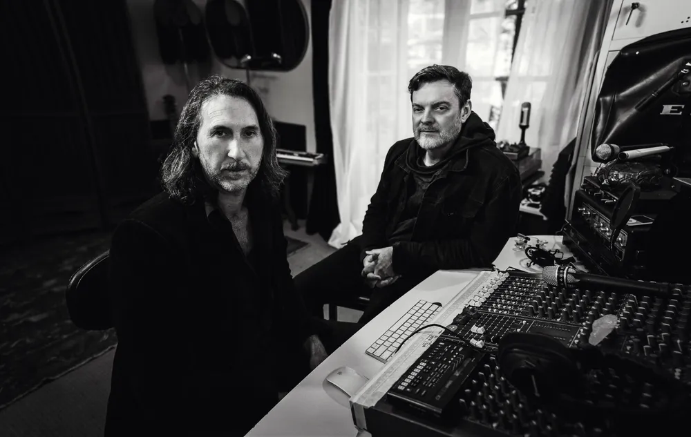 Black and white picture of two musicians in a recording studio sitting in front of a big mixer and other studio equipments.